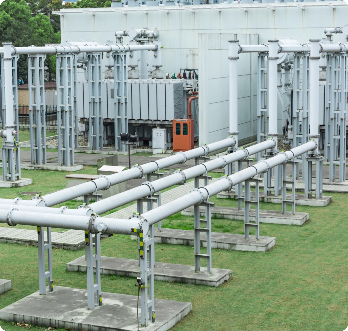 Industrial facility with metal pipelines and a large transformer, set against green grass and a gray building backdrop.